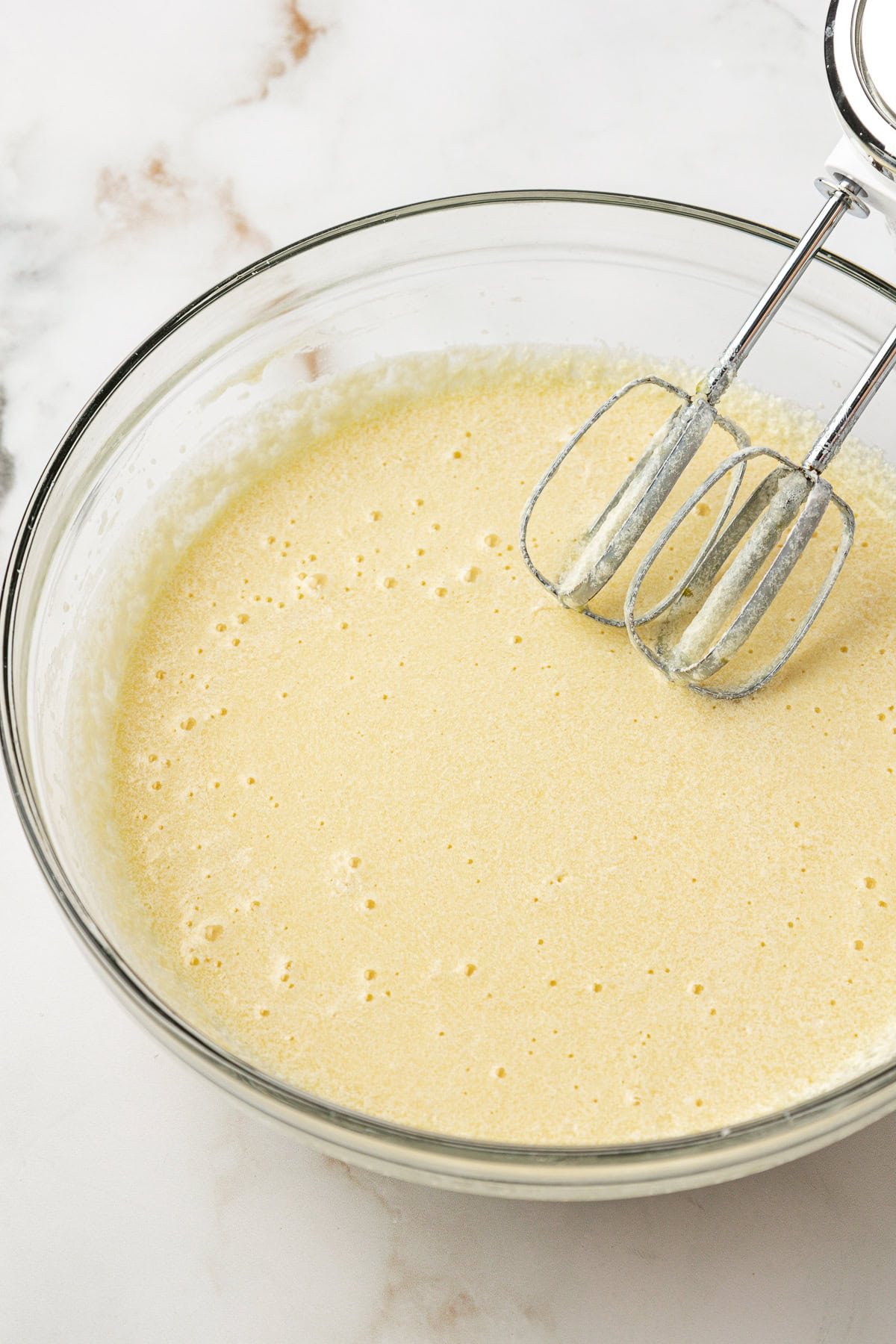 A glass bowl filled with a pale, frothy cake batter sits on a marble countertop. An electric hand mixer with two beaters rests on the edge of the bowl.