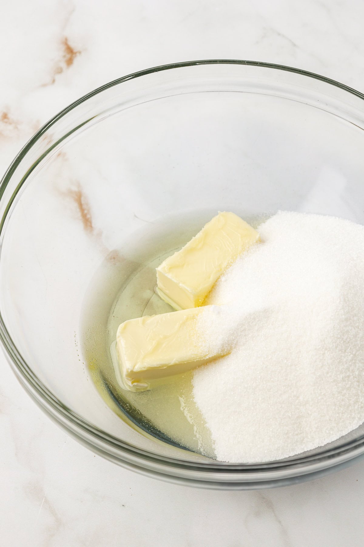 A glass bowl containing two sticks of butter and a pile of granulated white sugar on a marble surface.