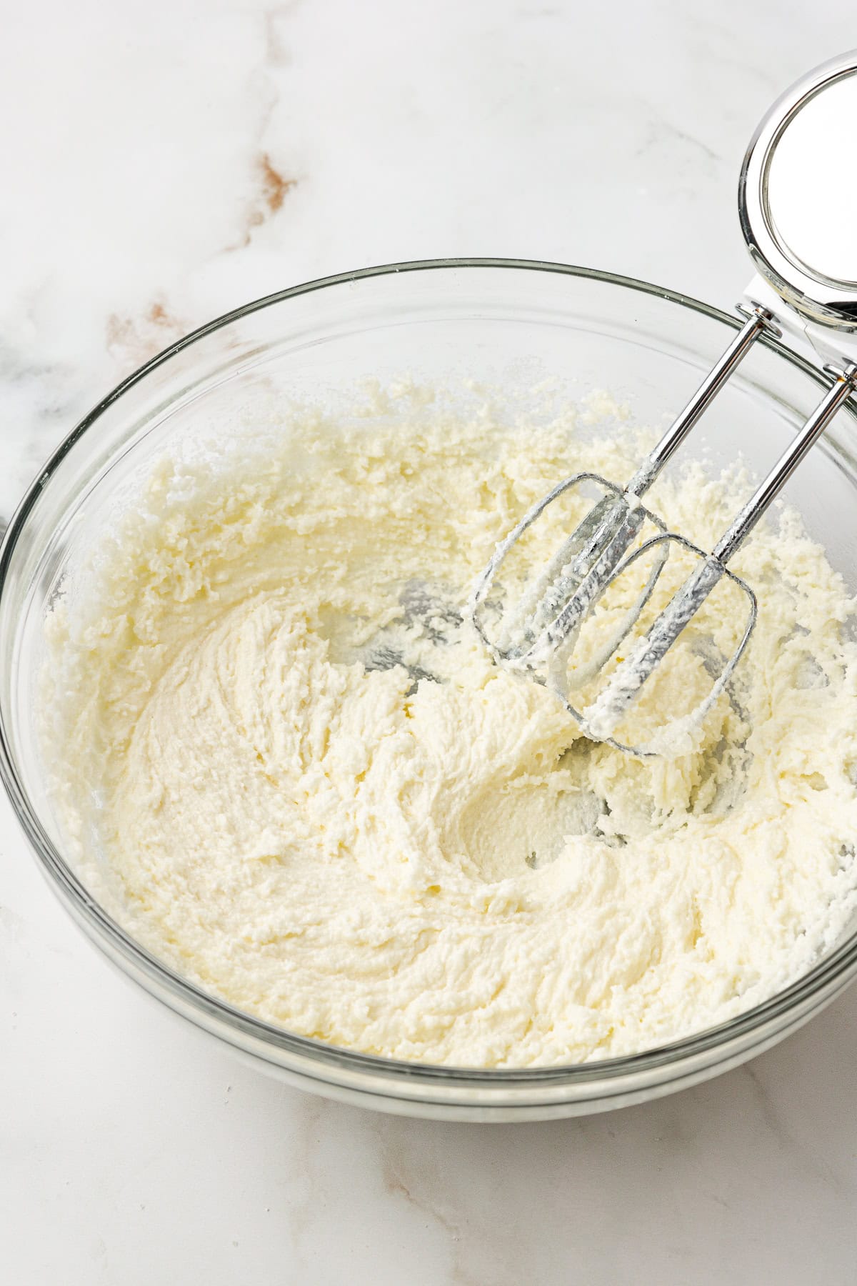 A glass bowl containing creamed butter and sugar with an electric hand mixer resting inside on a white marble surface.