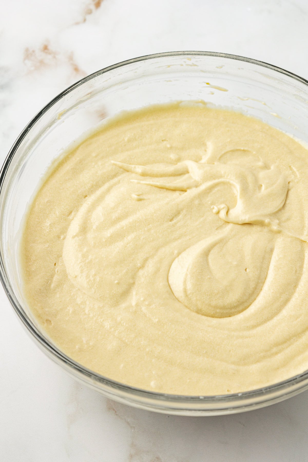A glass bowl filled with smooth, creamy cake batter sits on a white marble surface. The batter is light yellow and has visible swirls on the surface.