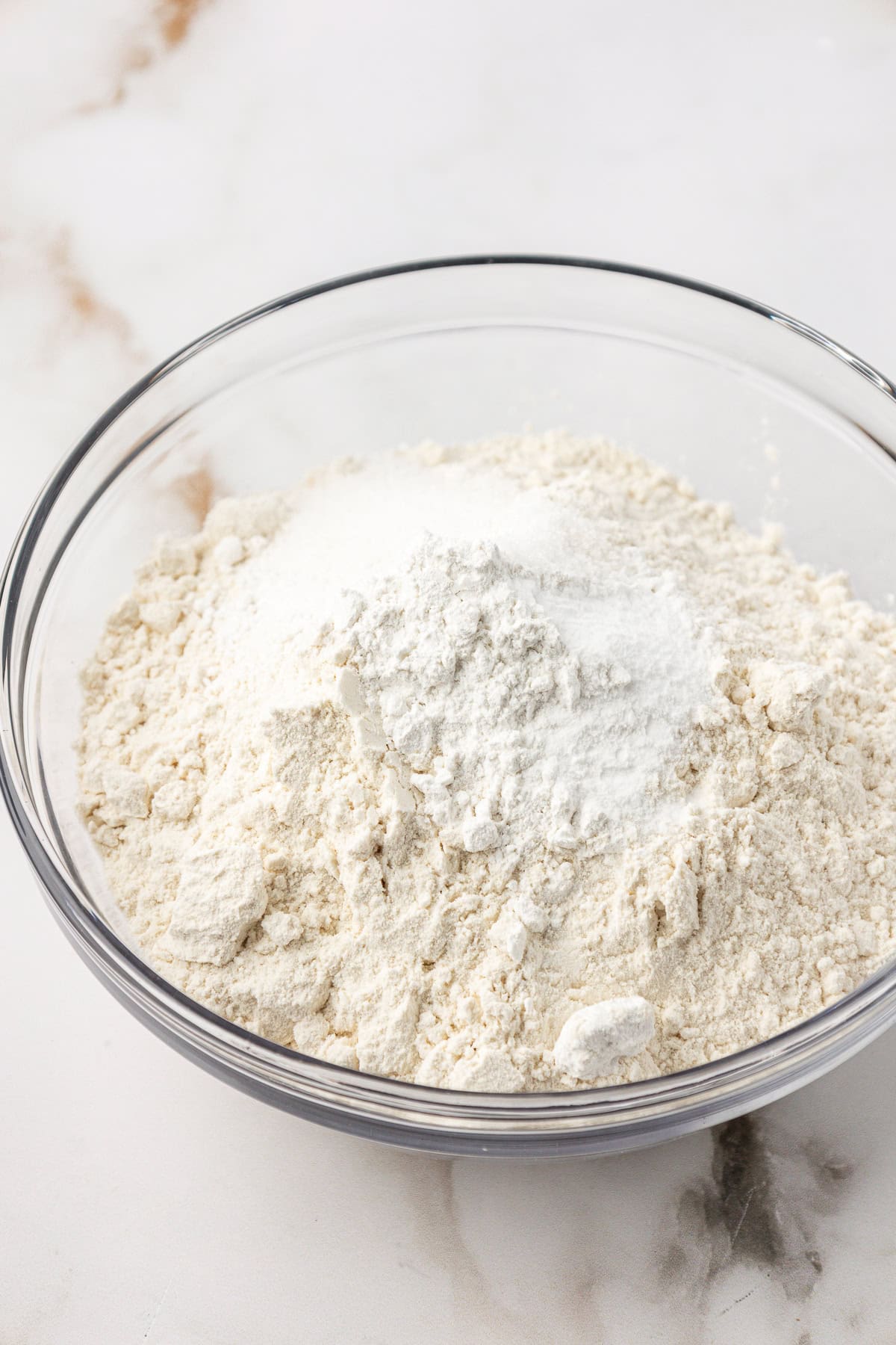 A clear glass bowl filled with a mixture of flour and white powder, likely baking soda or baking powder, sits on a white marble surface.