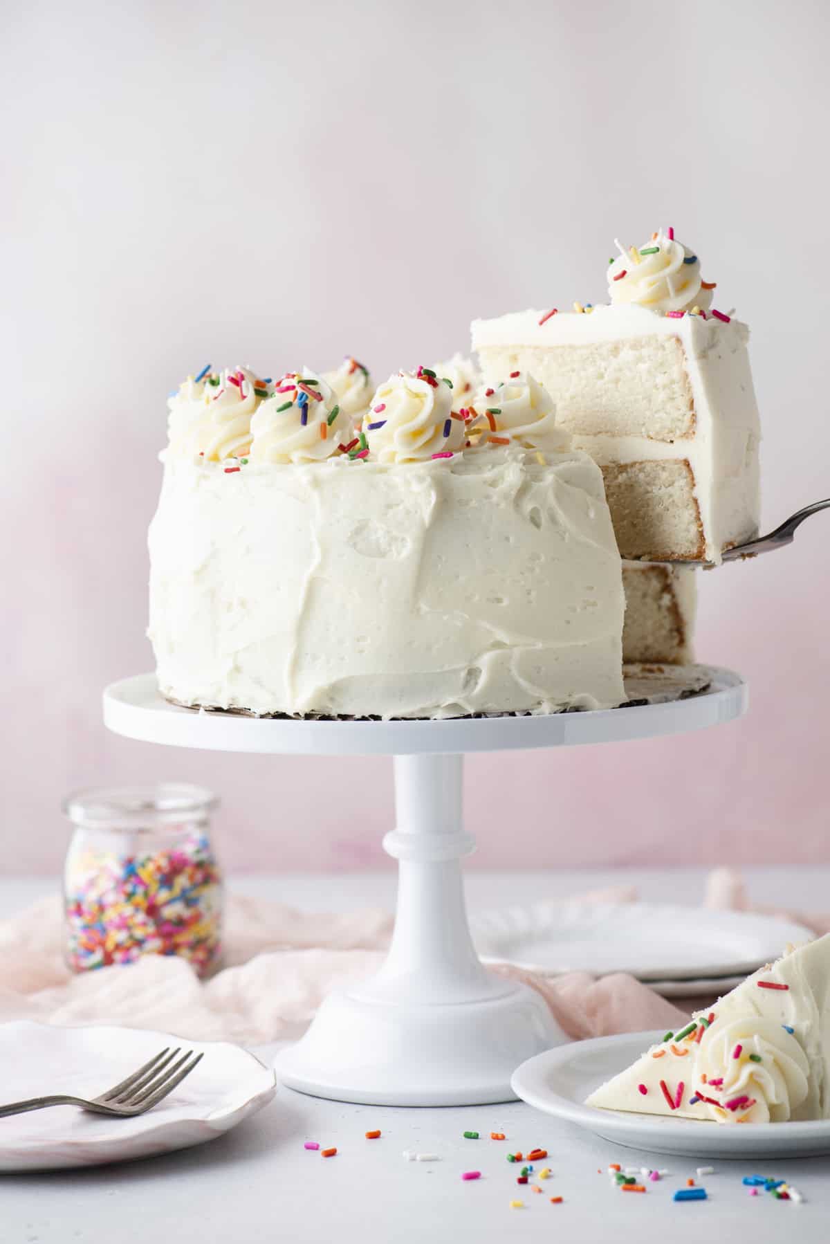 A white frosted layer cake with swirls of icing and colorful sprinkles sits on a cake stand. A slice is being lifted, revealing the cake’s fluffy interior. A jar of sprinkles, plates, and utensils are nearby.
