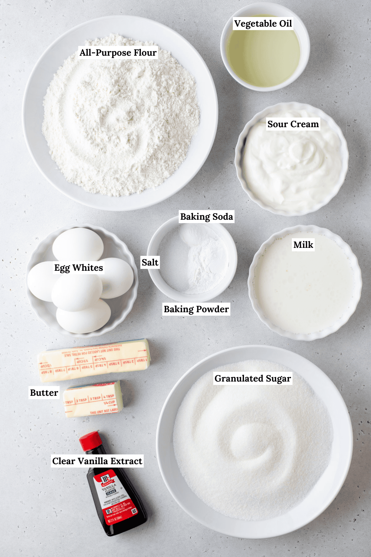 Top-down view of labeled baking ingredients on a white surface, including all-purpose flour, granulated sugar, butter sticks, eggs, vanilla extract, vegetable oil, sour cream, milk, baking powder, baking soda, and salt.
