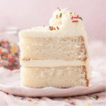 A close-up of a slice of white cake with two layers, filled and topped with white frosting and colorful sprinkles, on a light pink plate. A jar of sprinkles is blurred in the background.