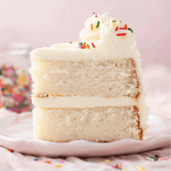 A close-up of a slice of white cake with two layers, filled and topped with white frosting and colorful sprinkles, on a light pink plate. A jar of sprinkles is blurred in the background.