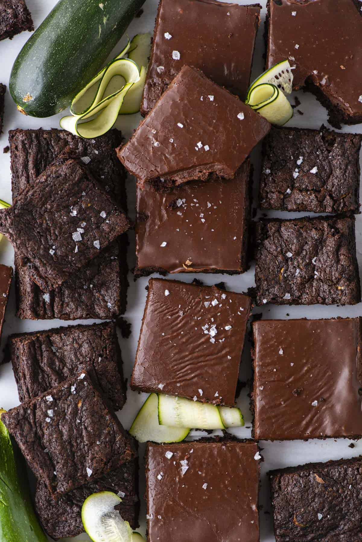 A top view of chocolate brownies, some with glossy frosting and a sprinkle of sea salt, arranged with fresh zucchini and zucchini ribbons on a white surface.