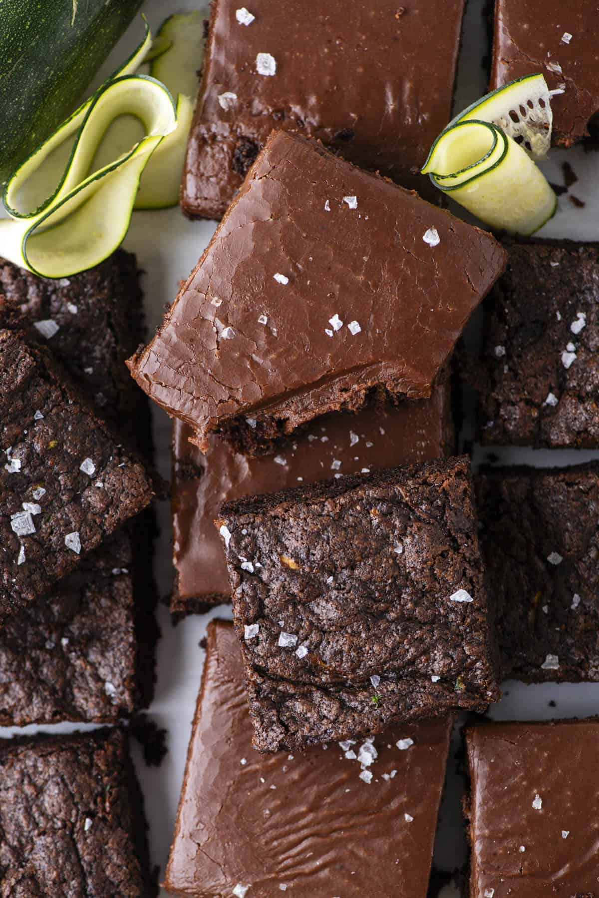 A close-up of chocolate brownies topped with flaky sea salt, some with chocolate frosting. Zucchini ribbons are placed in the corner, hinting at a possible ingredient in the brownies.