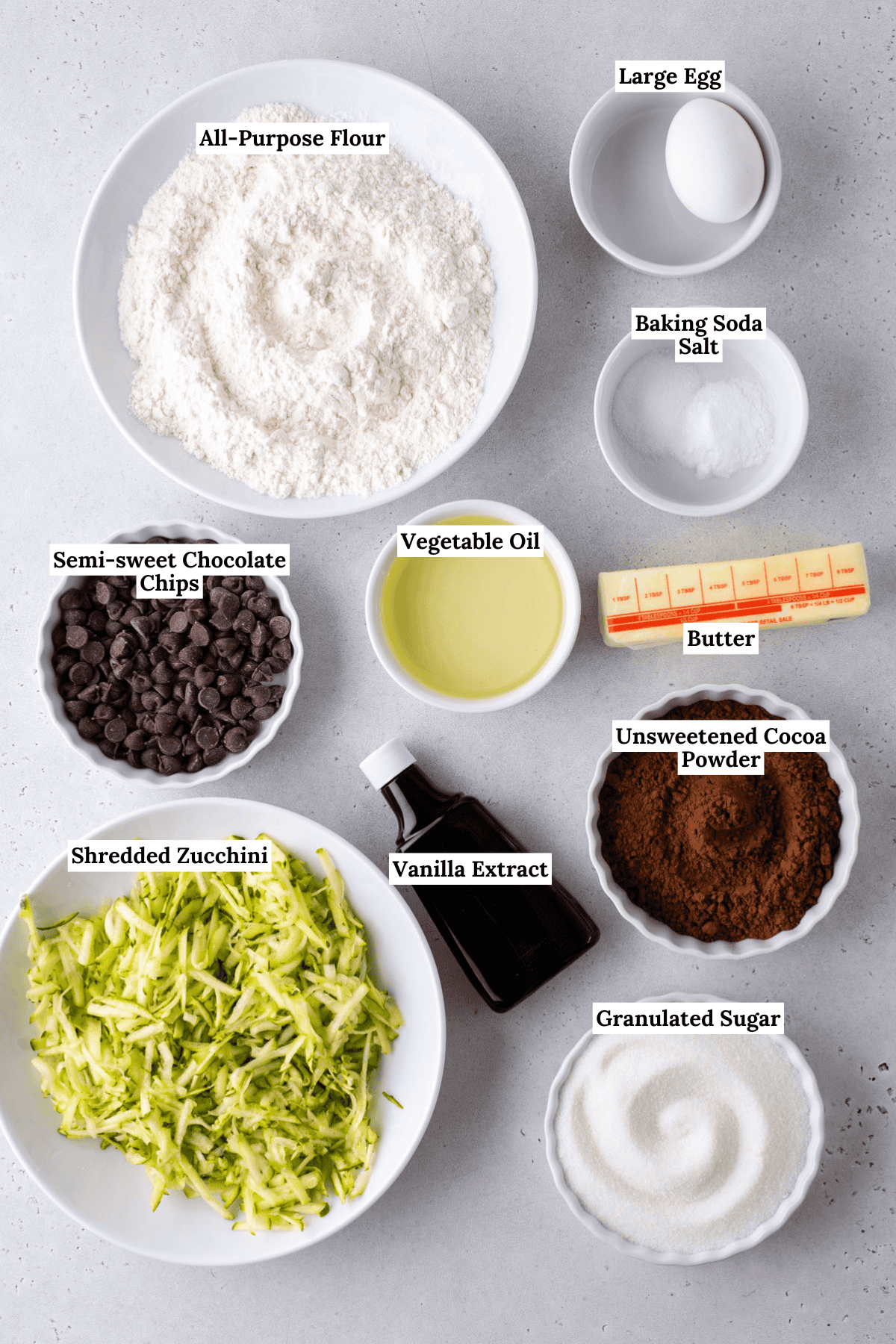 Top-down view of labeled baking ingredients in bowls and plates, including flour, egg, baking soda, salt, butter, cocoa powder, granulated sugar, vegetable oil, vanilla extract, shredded zucchini, and chocolate chips.