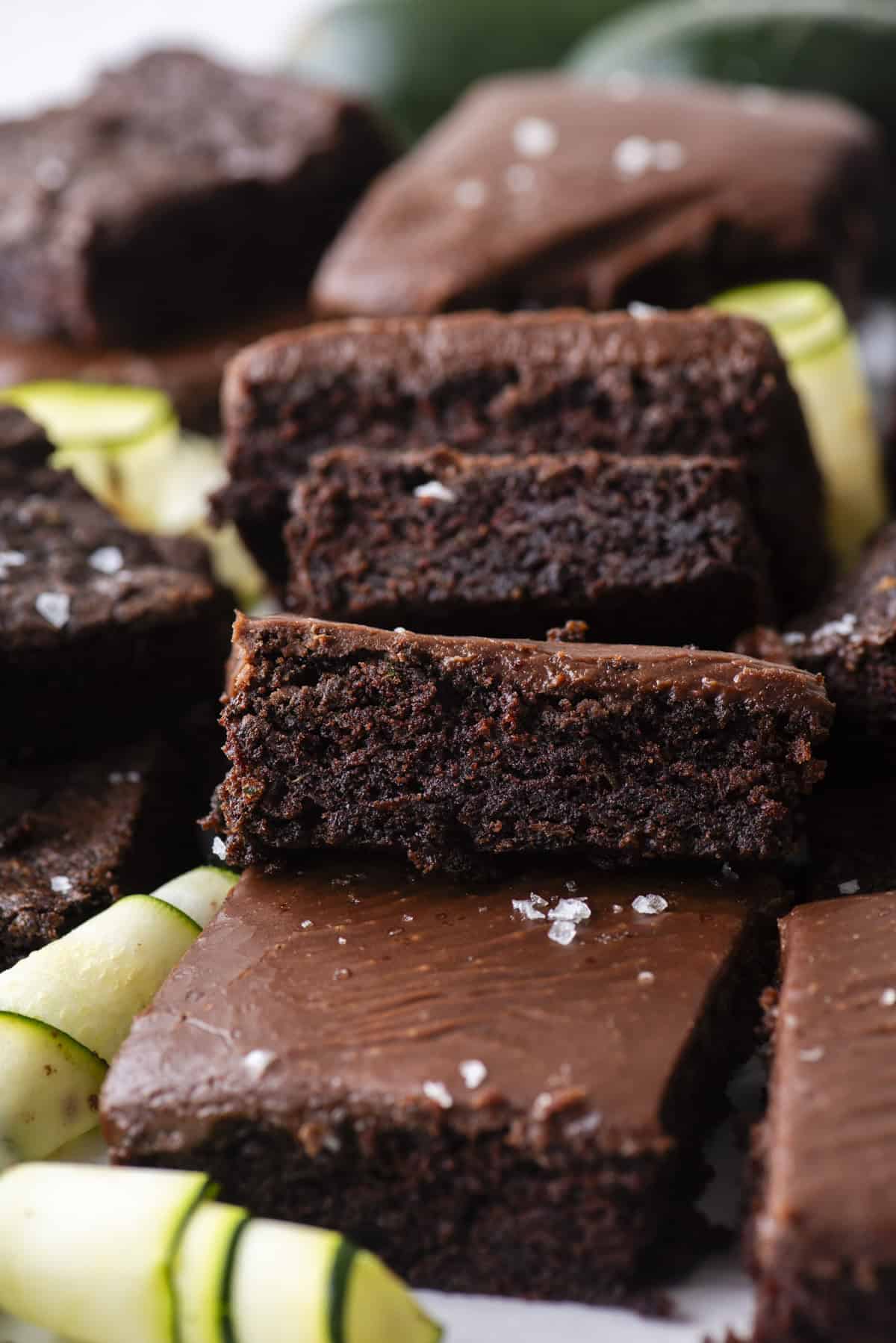 A close-up of rich, fudgy chocolate brownies stacked on top of each other, sprinkled with flaky sea salt. Slices of fresh zucchini are scattered around the brownies.