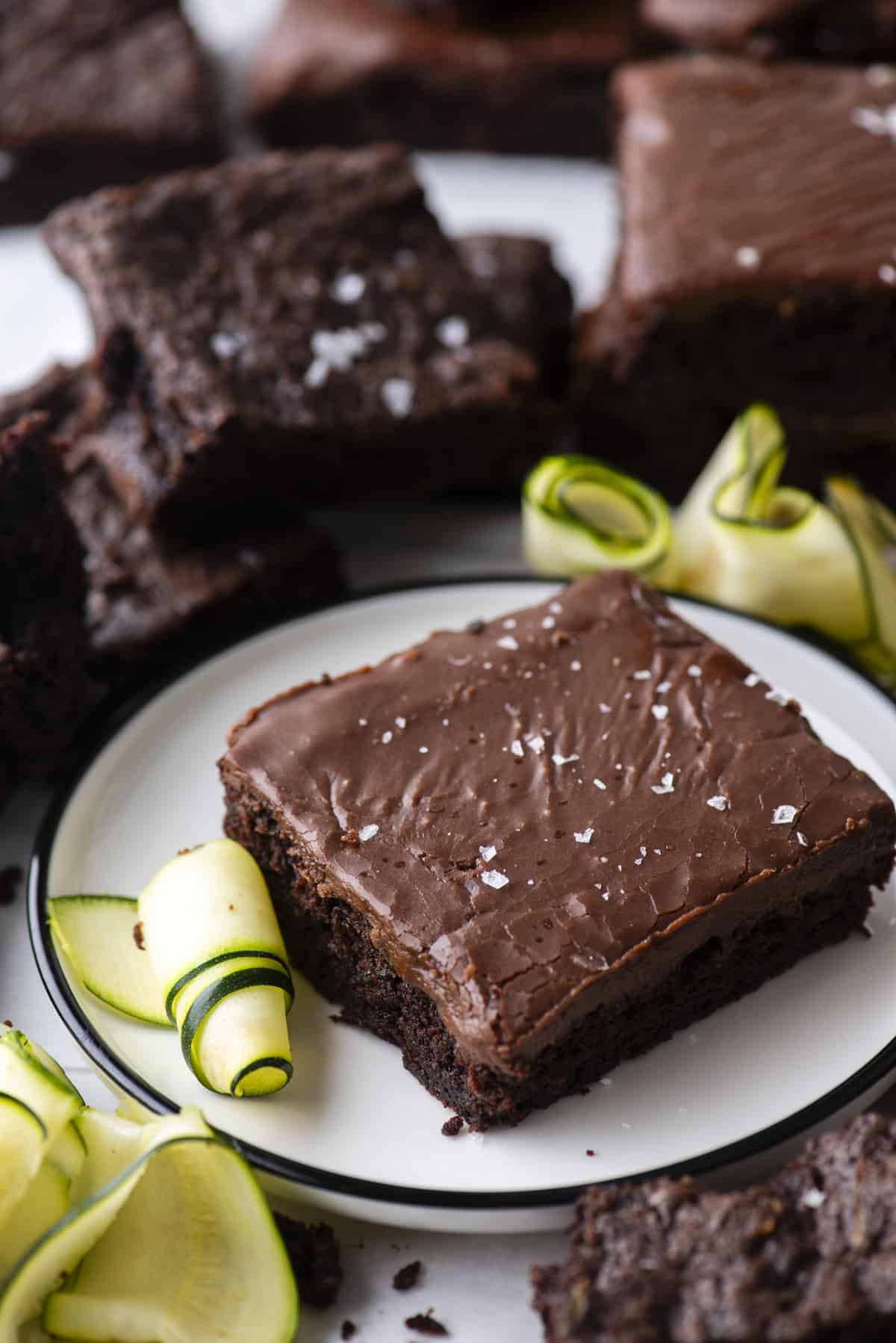 A chocolate brownie topped with flaky sea salt sits on a white plate, surrounded by chocolate brownies and garnished with fresh zucchini ribbons.