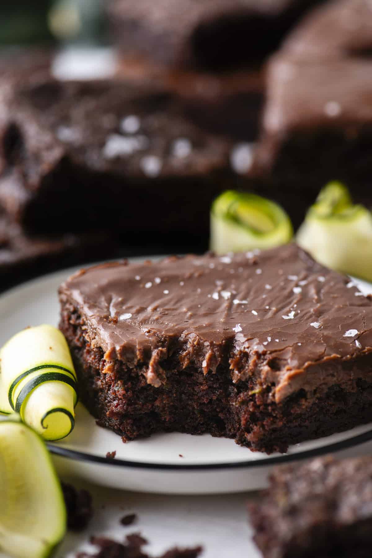 A close-up of a chocolate frosted brownie with a bite taken out, garnished with sea salt flakes and zucchini ribbons, served on a white plate with more brownies in the background.