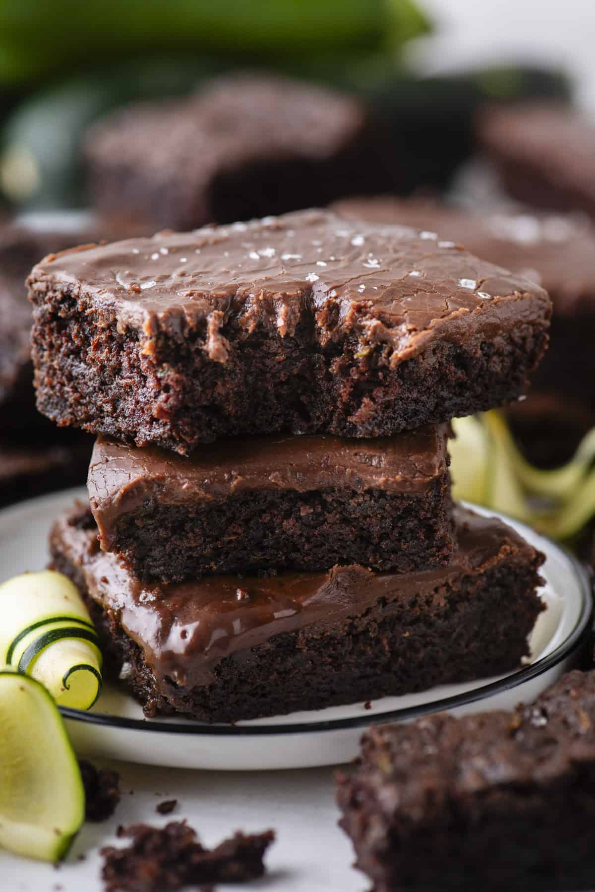 A stack of three chocolate brownies with glossy frosting, sprinkled with sea salt, sits on a white plate. Slices of zucchini are beside the brownies. The background shows more brownies.