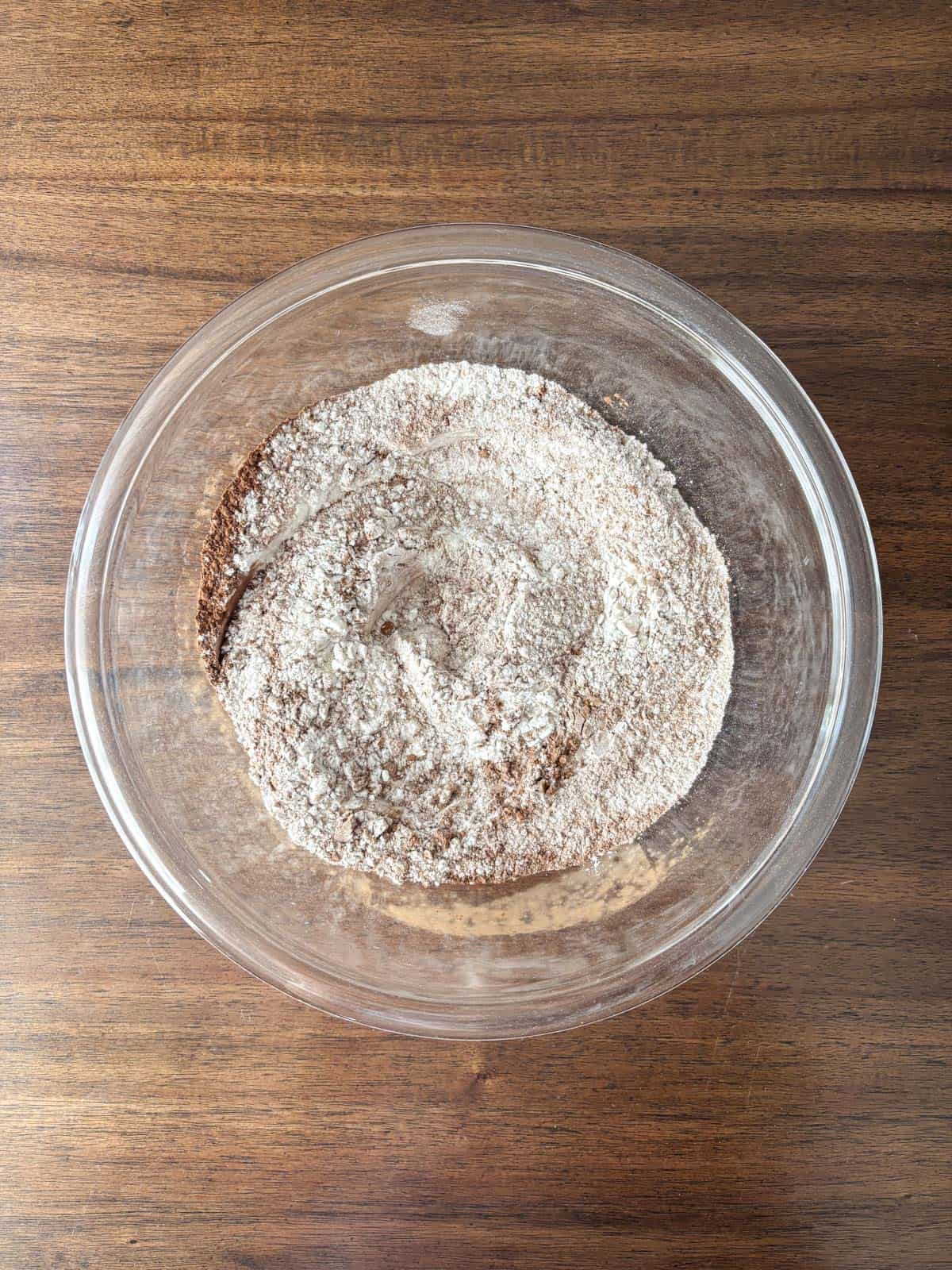 A glass bowl filled with a mixture of dry ingredients, including flour and cocoa powder, sits on a wooden surface.