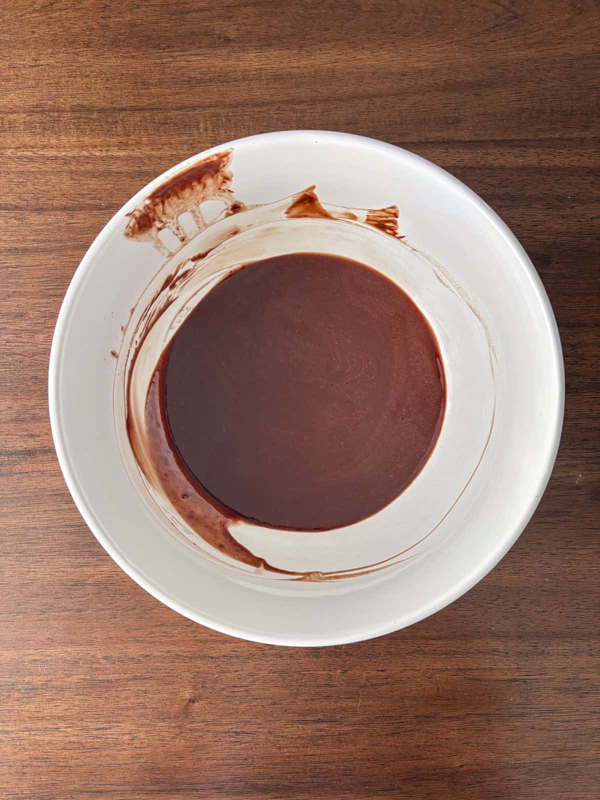 A white mixing bowl containing smooth, dark chocolate batter sits on a wooden surface. Some batter is smeared along the inner sides of the bowl.