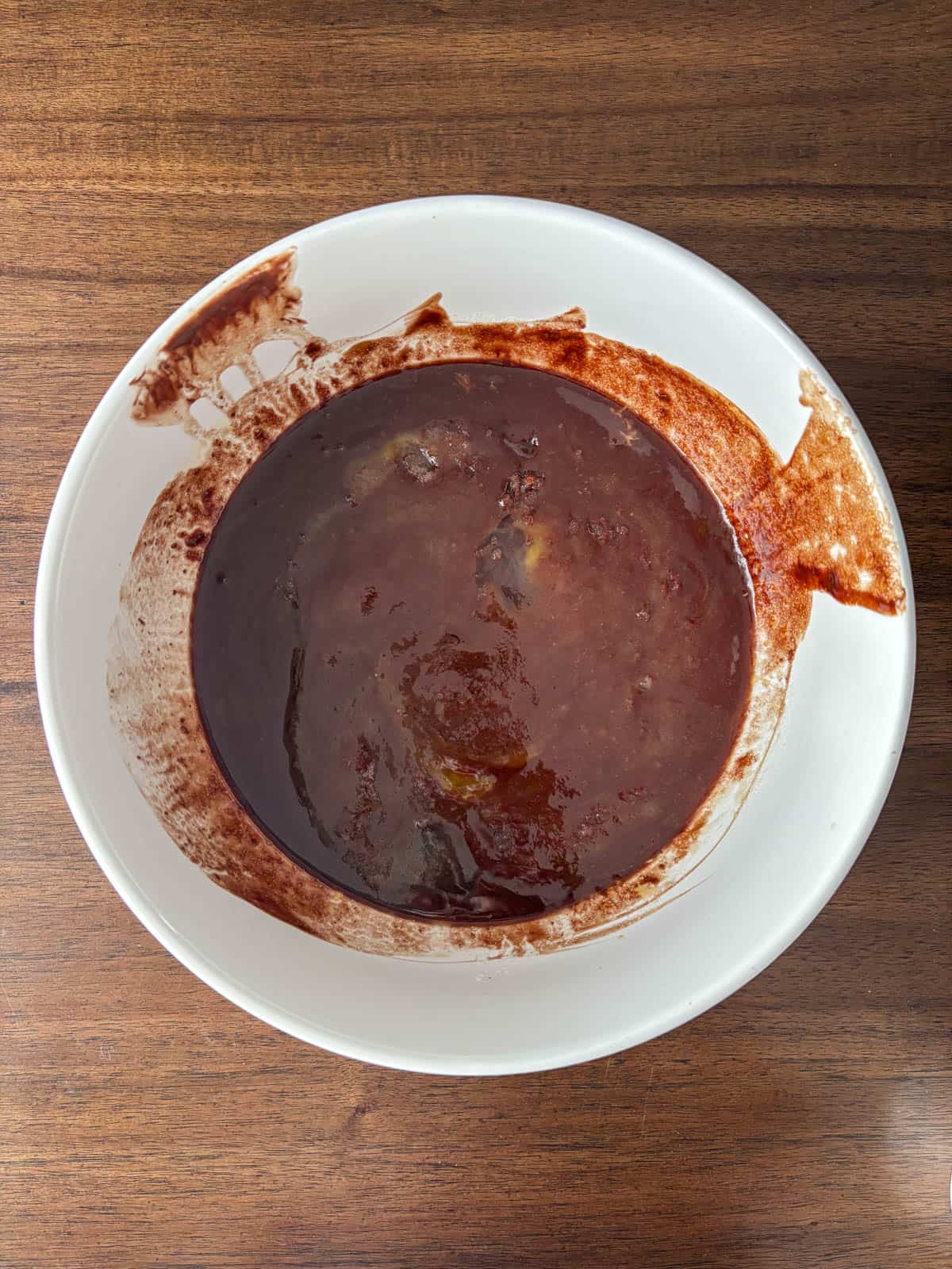A white bowl filled with dark brown, thick soup or stew sits on a wooden table, with some of the liquid splashed on the rim and side of the bowl.