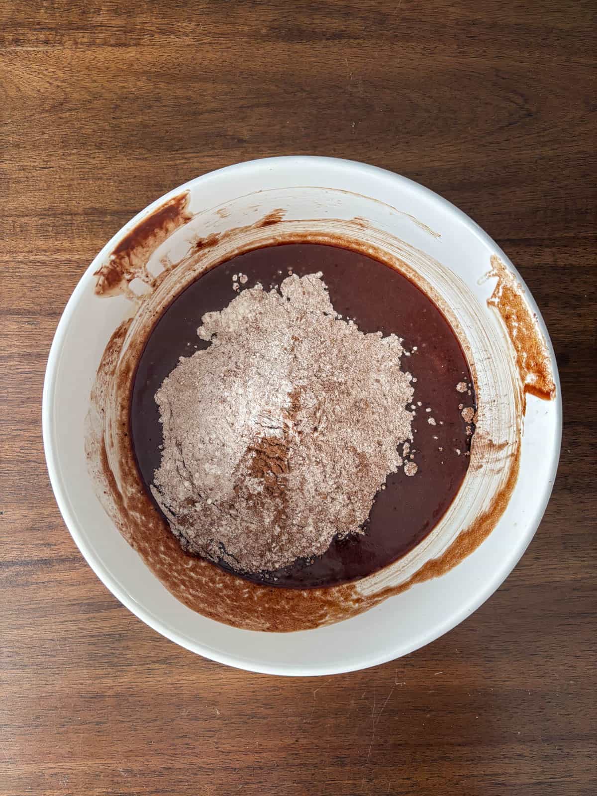 A white bowl on a wooden surface contains brownie batter with a mound of dry cocoa powder and flour in the center, partially mixed into the wet ingredients.