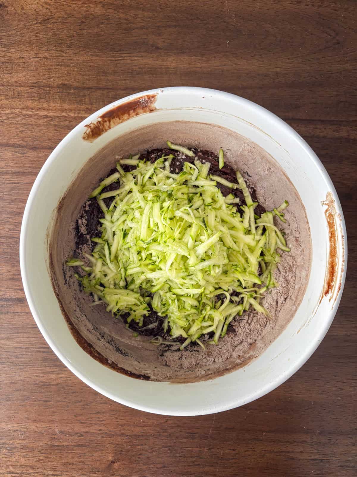 A white mixing bowl with chocolate cake batter and a pile of freshly grated zucchini on top, sitting on a wooden surface.