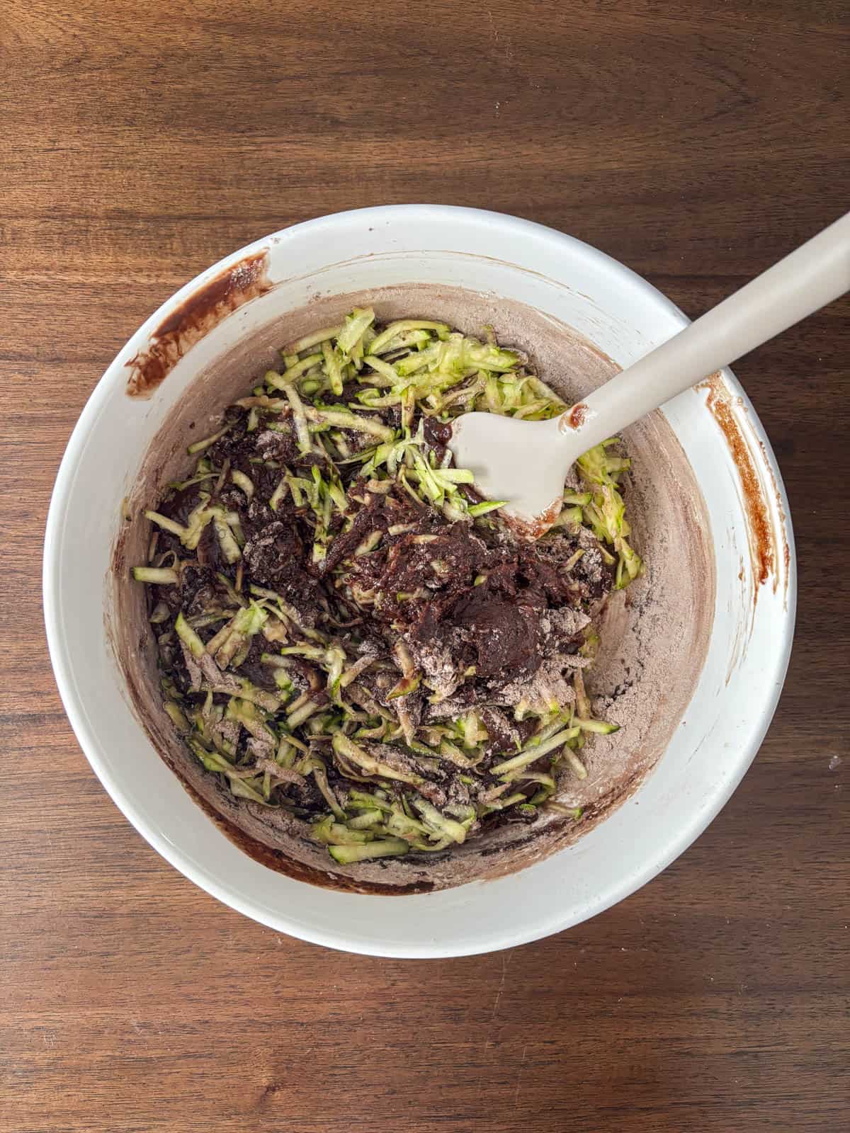 A white mixing bowl filled with brownie batter and shredded zucchini sits on a wooden surface. A white spatula is resting in the bowl, ready to stir the ingredients together.