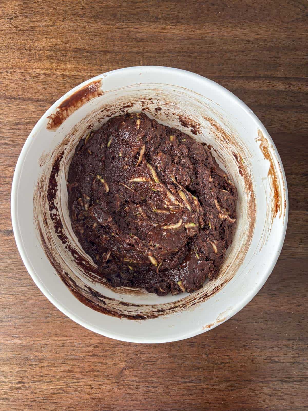 A white mixing bowl on a wooden surface contains thick, dark chocolate batter with visible shreds of zucchini mixed in.