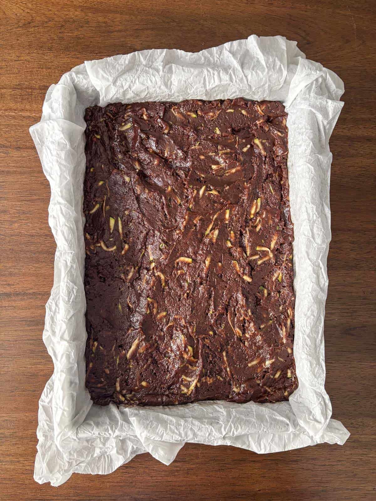 A rectangular baking pan lined with crinkled parchment paper holds unbaked chocolate brownie batter with visible almond slivers, placed on a wooden surface.