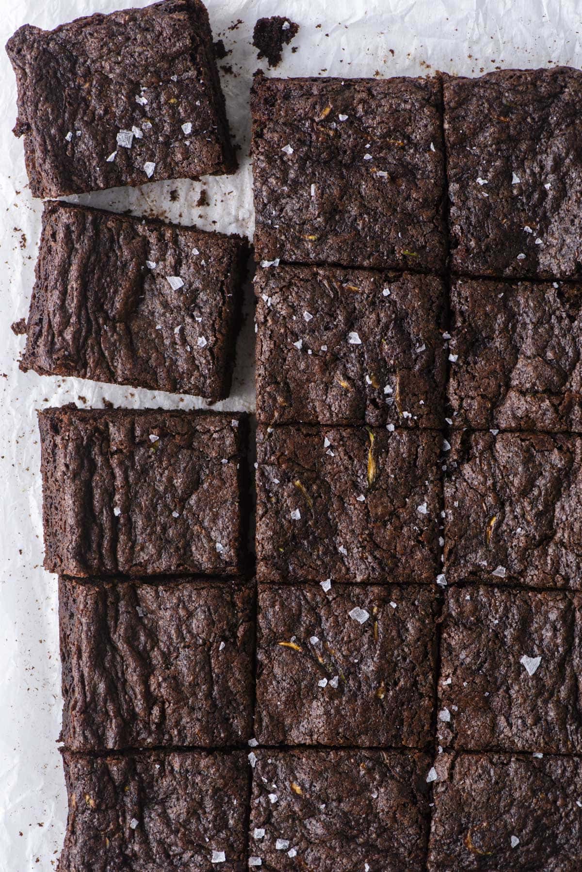A batch of chocolate brownies cut into squares on parchment paper. Two brownies are stacked in the top left corner. The brownies are topped with a sprinkle of flaky sea salt.