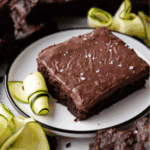 A chocolate zucchini brownie topped with flaky sea salt is served on a white plate, garnished with thinly sliced zucchini ribbons. Additional brownies and zucchini slices are visible in the background.