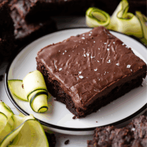 A chocolate zucchini brownie topped with flaky sea salt is served on a white plate, garnished with thinly sliced zucchini ribbons. Additional brownies and zucchini slices are visible in the background.