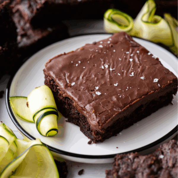 A chocolate zucchini brownie topped with flaky sea salt is served on a white plate, garnished with thinly sliced zucchini ribbons. Additional brownies and zucchini slices are visible in the background.
