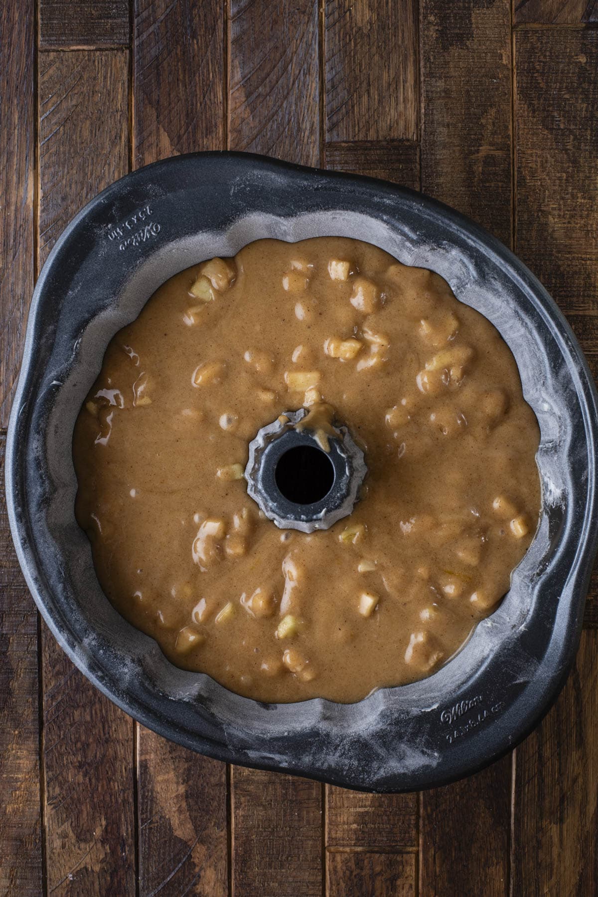 A bundt cake pan filled with light brown batter containing small apple chunks, sitting on a dark wooden surface. The pan is floured, and the batter is ready to be baked.