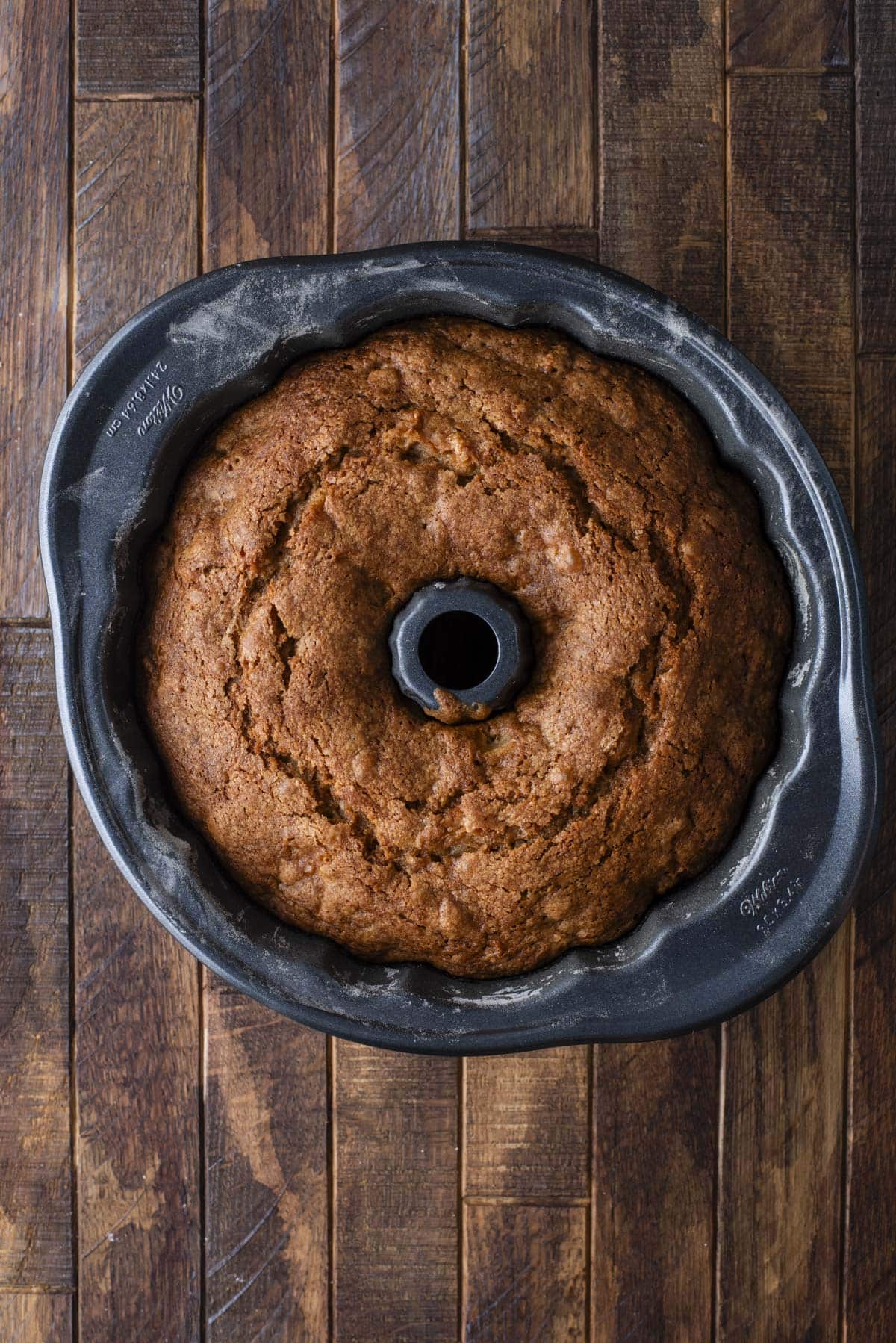 A golden-brown bundt cake sits freshly baked in a dark bundt pan on a wooden surface, viewed from above.