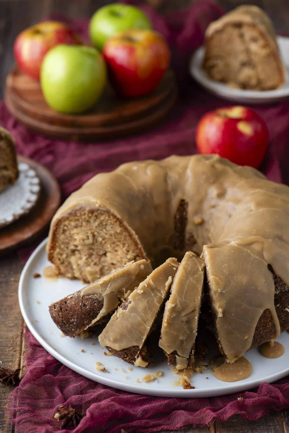 A glazed apple bundt cake with several slices cut, displayed on a white plate. Fresh red and green apples are in the background, along with more cake on a small plate, all set on a rustic wooden table with a burgundy cloth.