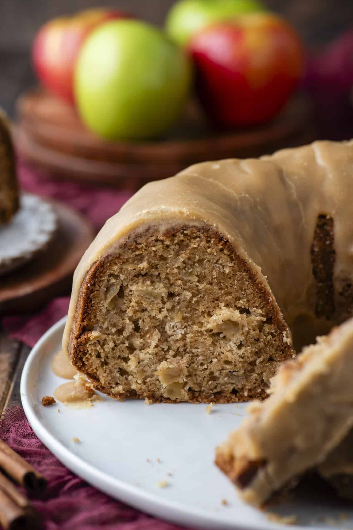 A close-up of a sliced bundt cake with caramel glaze, showing a moist, dense interior. In the background, green and red apples are slightly out of focus on a wooden plate.