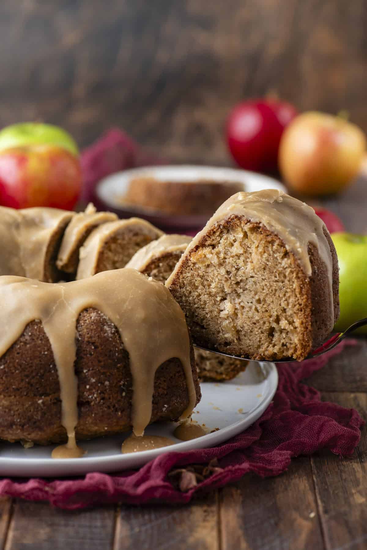 A sliced bundt cake with caramel glaze sits on a white plate, surrounded by fresh apples. A cake server lifts a piece, showing the moist interior. The background features a rustic wooden surface and a burgundy cloth.
