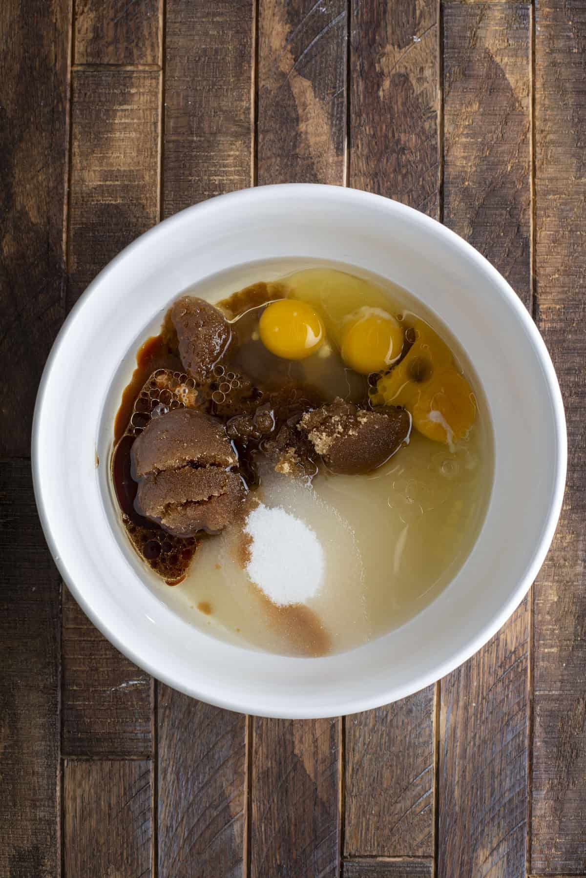 A white bowl on a wooden surface contains brown sugar, white sugar, eggs, vanilla extract, and liquid ingredients, ready to be mixed for baking.