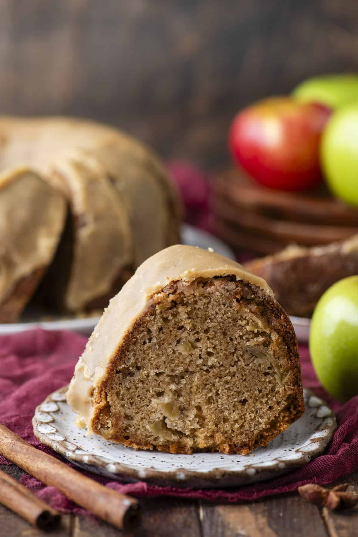 A slice of glazed bundt cake with visible apple pieces sits on a plate, with fresh red and green apples and cinnamon sticks nearby. The rest of the cake and a dark rustic background are blurred behind.