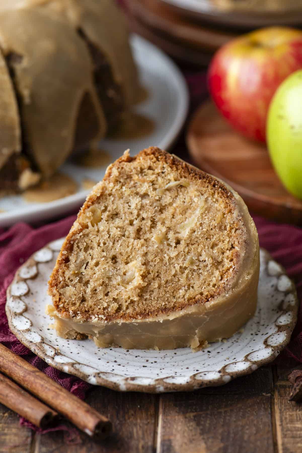 A slice of glazed apple cake sits on a ceramic plate, showing a moist, spiced interior. Whole apples and cinnamon sticks are nearby, and the rest of the cake is in the background.