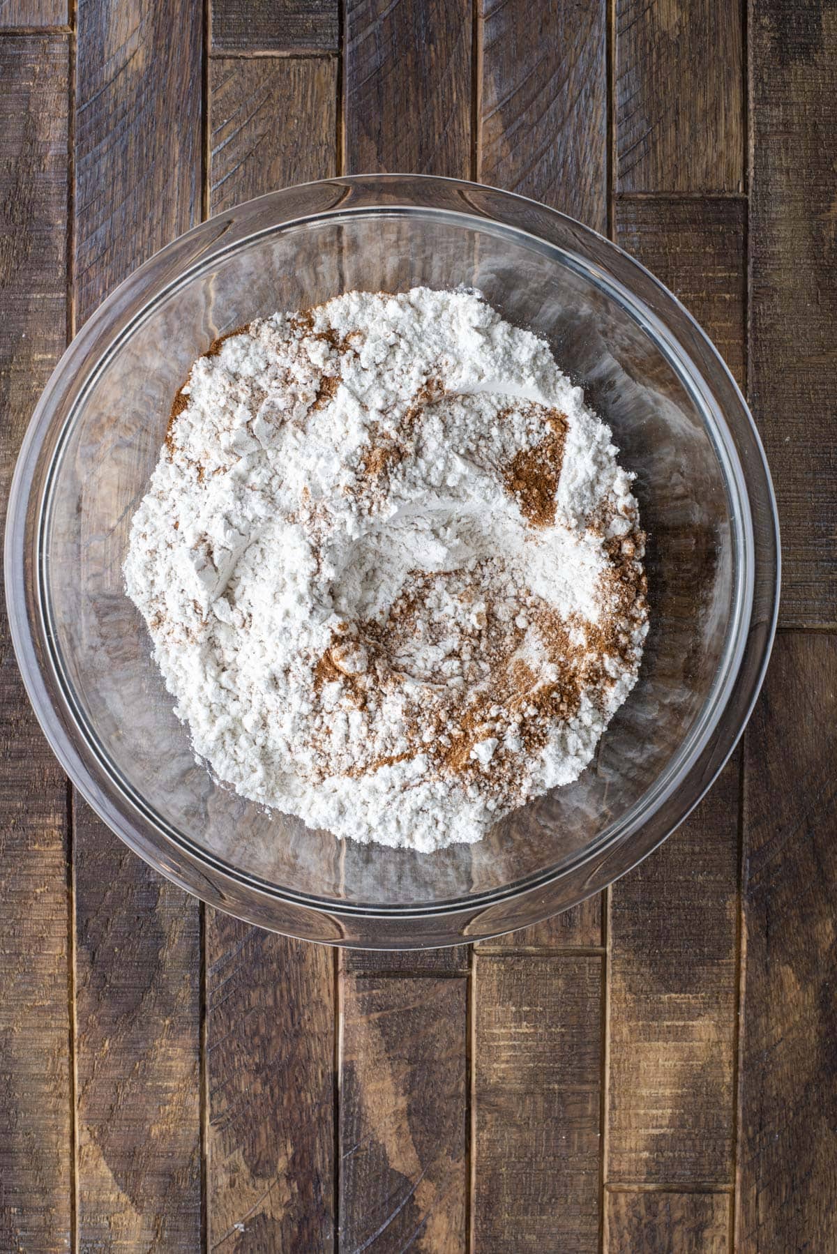A glass bowl filled with flour and cinnamon sits on a wooden surface. The ingredients are unmixed and arranged in a mound in the center of the bowl.