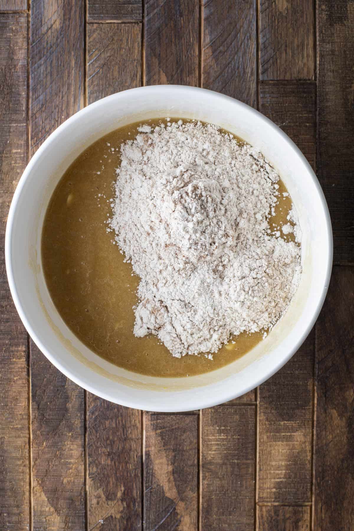 A white bowl filled with a light brown liquid mixture and topped with a mound of flour, sitting on a rustic wooden surface.