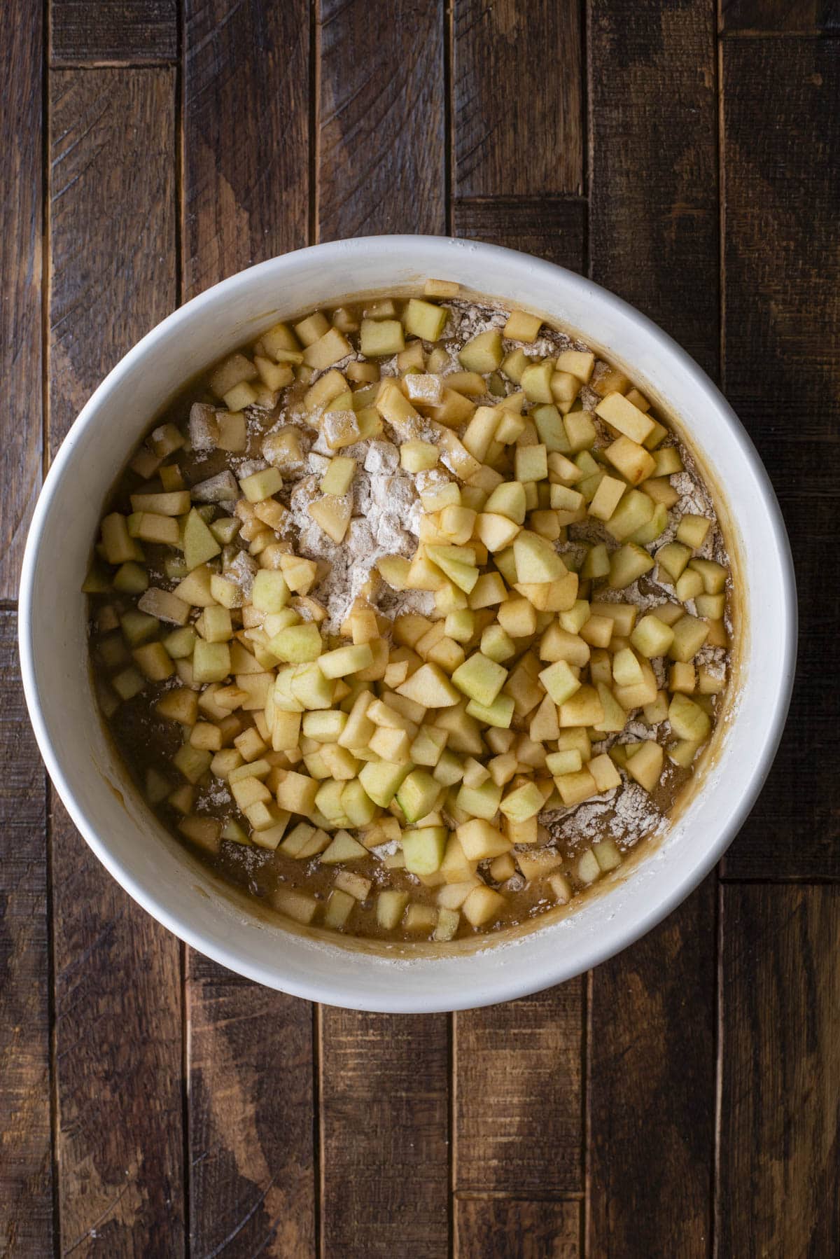A white mixing bowl filled with diced apples, flour, and batter sits on a rustic wooden surface, ready to be mixed for baking.