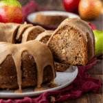 A close-up of a glazed bundt cake with a slice being lifted from the whole cake. The cake is on a white plate with a red cloth underneath, and apples are visible in the background.