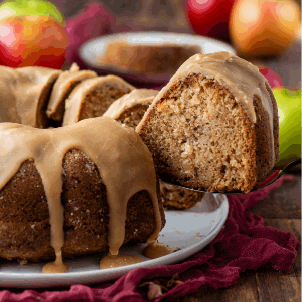 A close-up of a glazed bundt cake with a slice being lifted from the whole cake. The cake is on a white plate with a red cloth underneath, and apples are visible in the background.