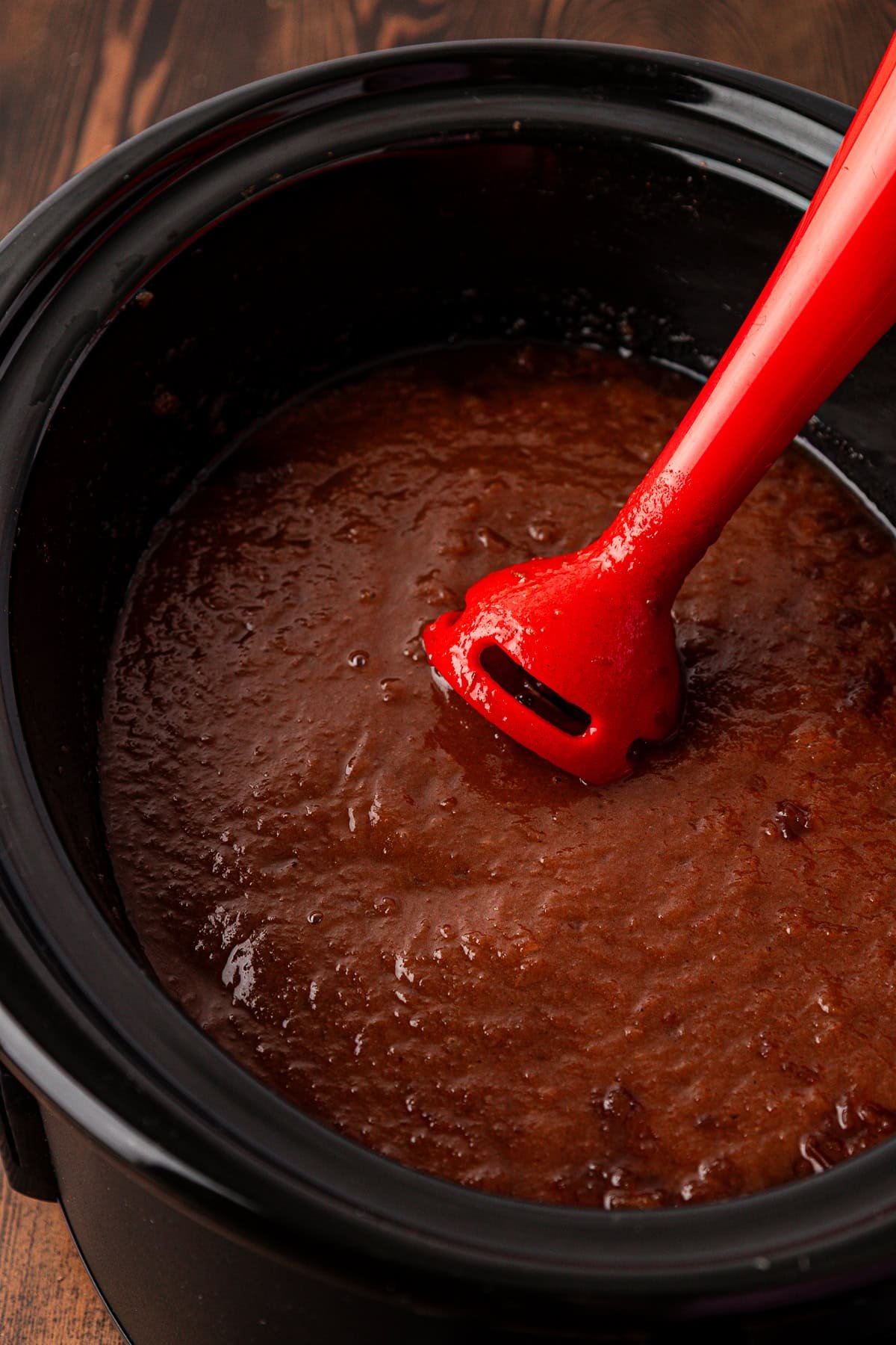 A red immersion blender is blending a thick, brown mixture inside a black slow cooker, placed on a wooden surface.
