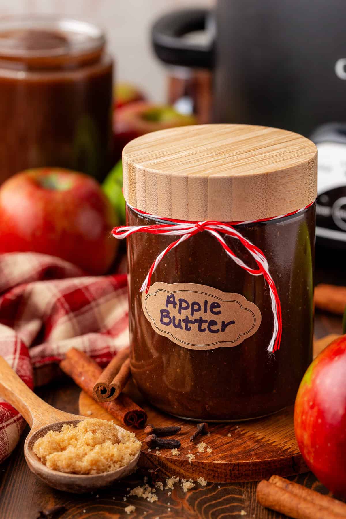 A glass jar labeled Apple Butter with a wooden lid and red-and-white string sits on a table with fresh apples, cinnamon sticks, brown sugar, and a red plaid cloth.