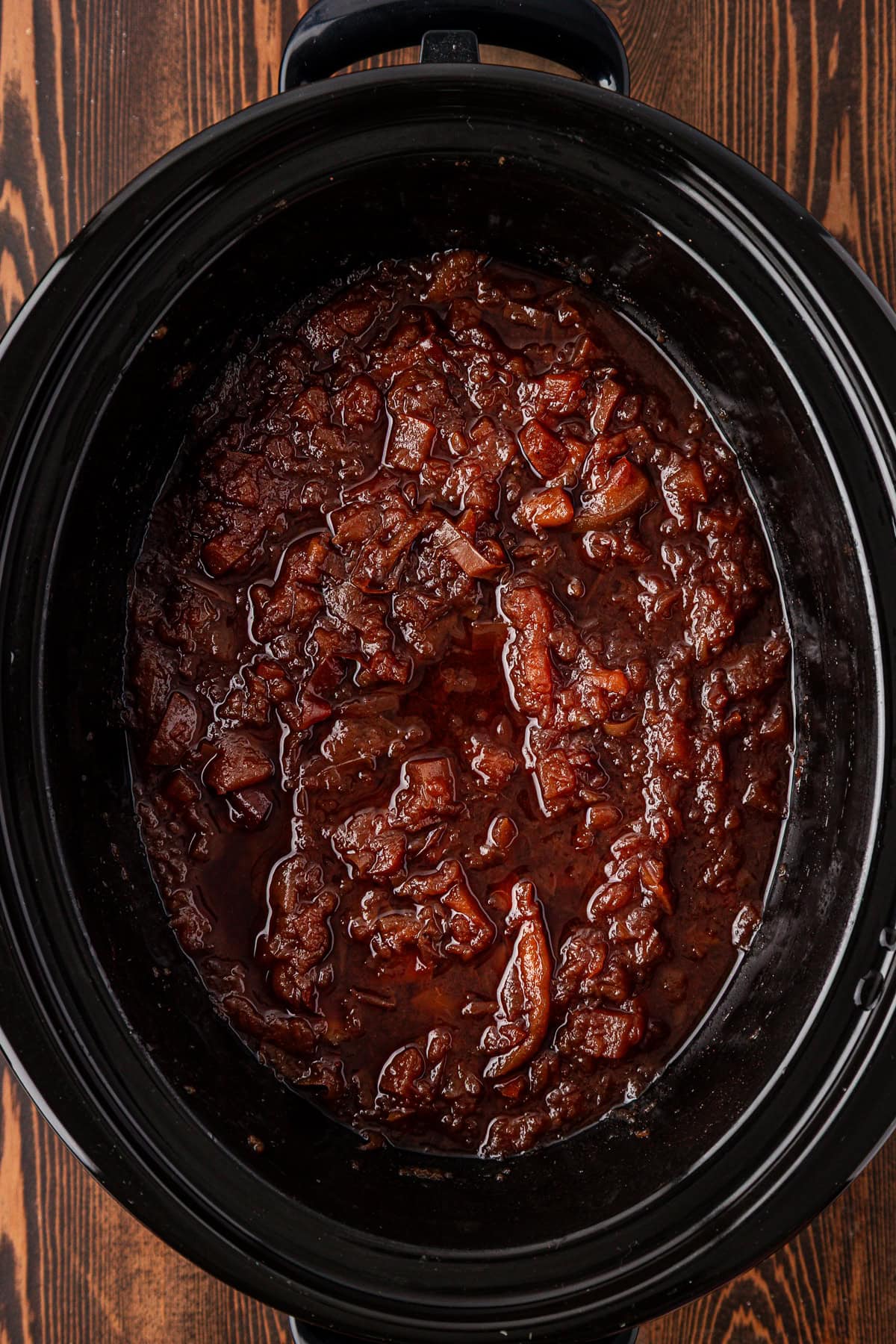 A slow cooker filled with thick, dark brown caramelized onions, showing a rich and glossy texture, sits on a wooden surface.