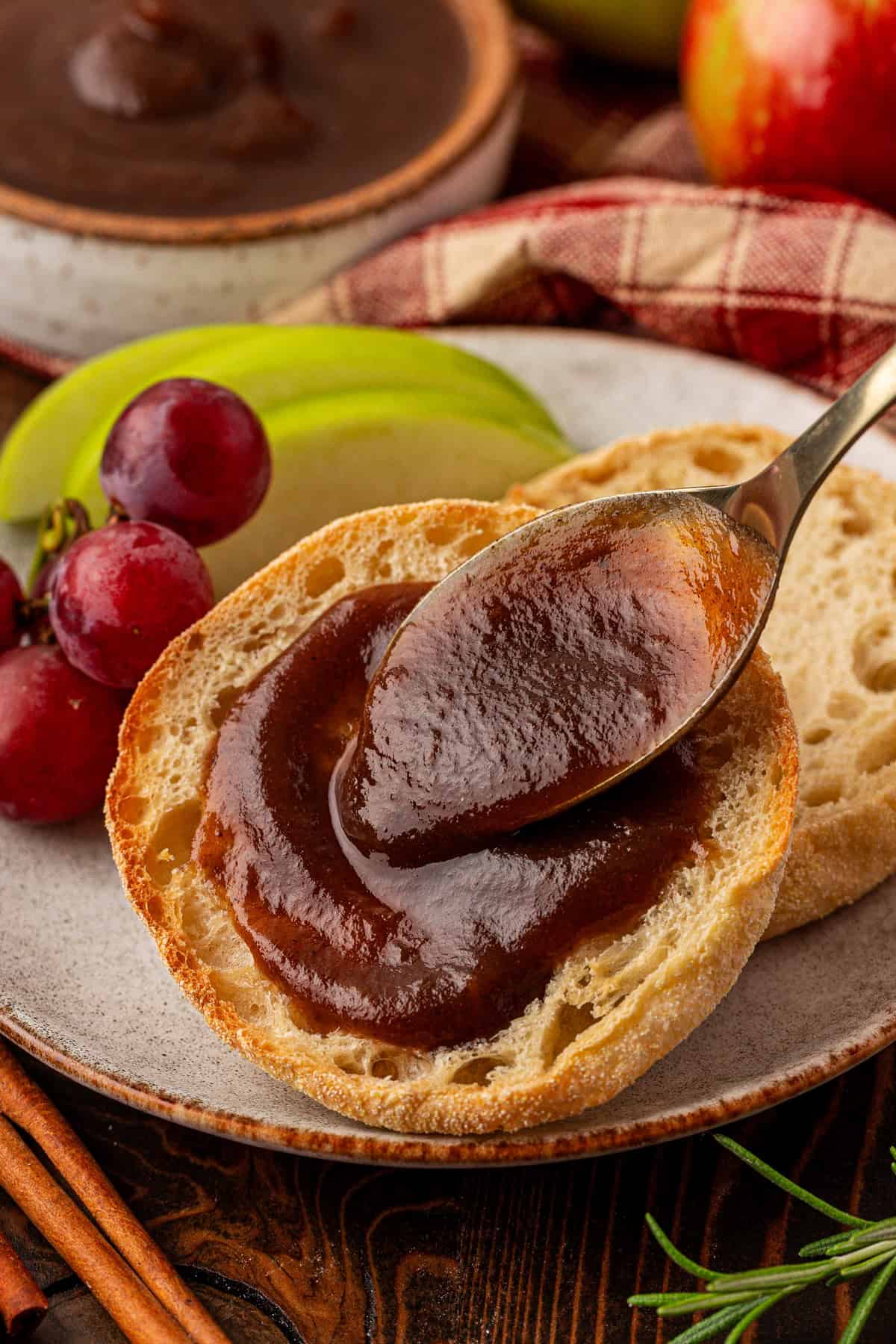 A spoon spreads thick apple butter on an English muffin half, with grapes, green apple slices, and a bowl of apple butter in the background, all arranged on a rustic plate.
