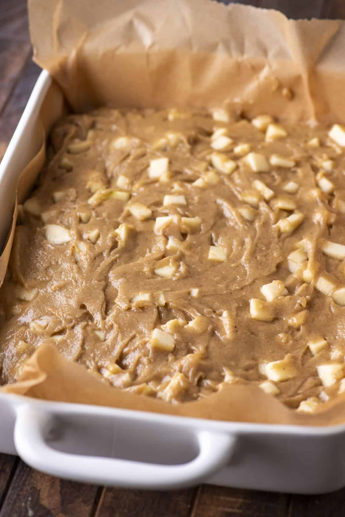 A white baking dish lined with brown parchment paper is filled with unbaked apple cake batter, showing chunks of fresh apple mixed throughout. The dish sits on a dark wooden surface.