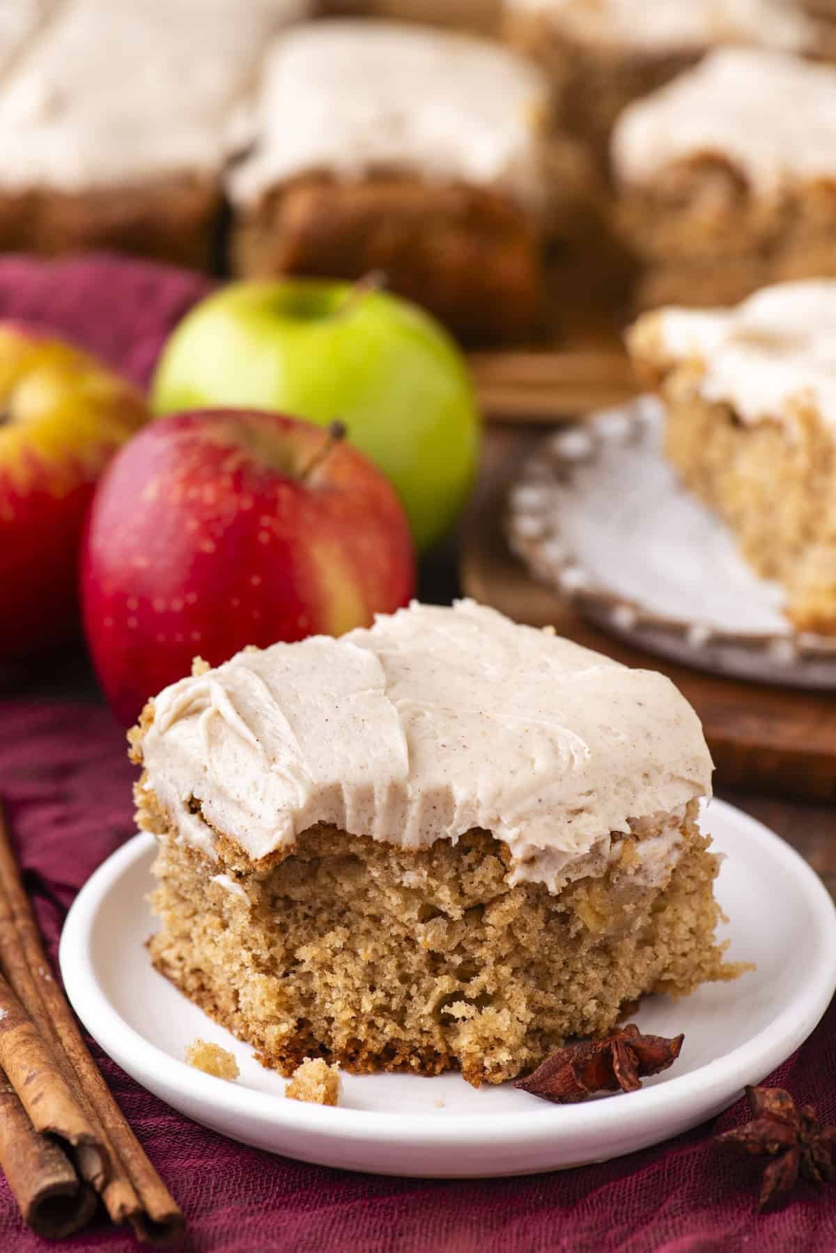 A square piece of spiced cake with creamy frosting sits on a white plate, surrounded by whole red and green apples, cinnamon sticks, and more frosted cake slices in the background.