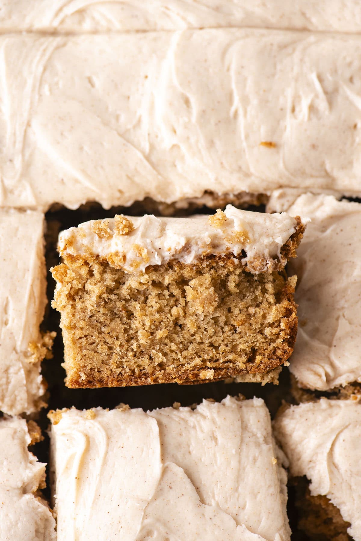 A close-up of a slice of moist, spiced cake with creamy frosting, lifted slightly above a tray of similar frosted cake pieces. The cake appears soft and dense with a thick layer of frosting on top.