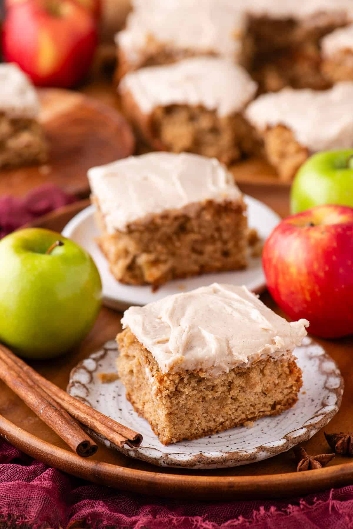 A close-up of frosted apple spice cake squares on plates, surrounded by fresh red and green apples and cinnamon sticks on a wooden tray. More cake pieces are visible in the background.