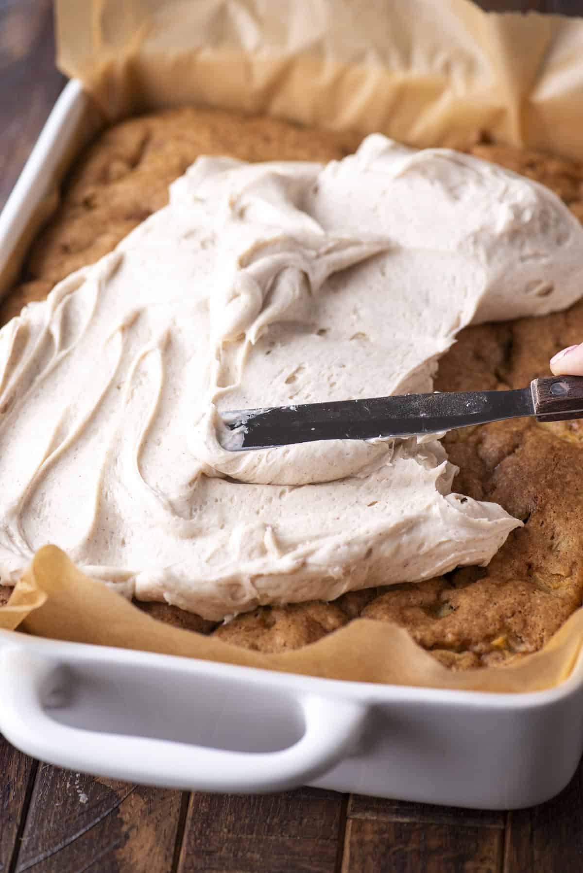 A hand spreads thick, creamy frosting over a large sheet cake in a white baking dish lined with parchment paper.